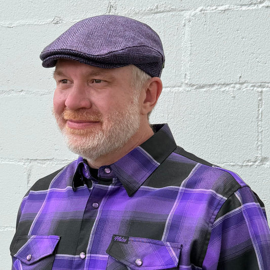 A man wearing a purple and black plaid button-down shirt and a matching purple flat cap, standing against a white painted brick wall.