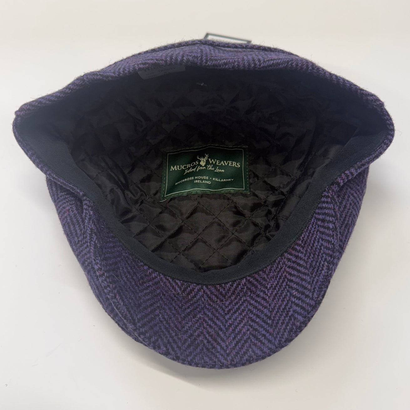 A top-down view of a purple herringbone tweed flat cap with a black quilted interior lining. A green 'Mick & Weavers' brand label is visible inside the crown.