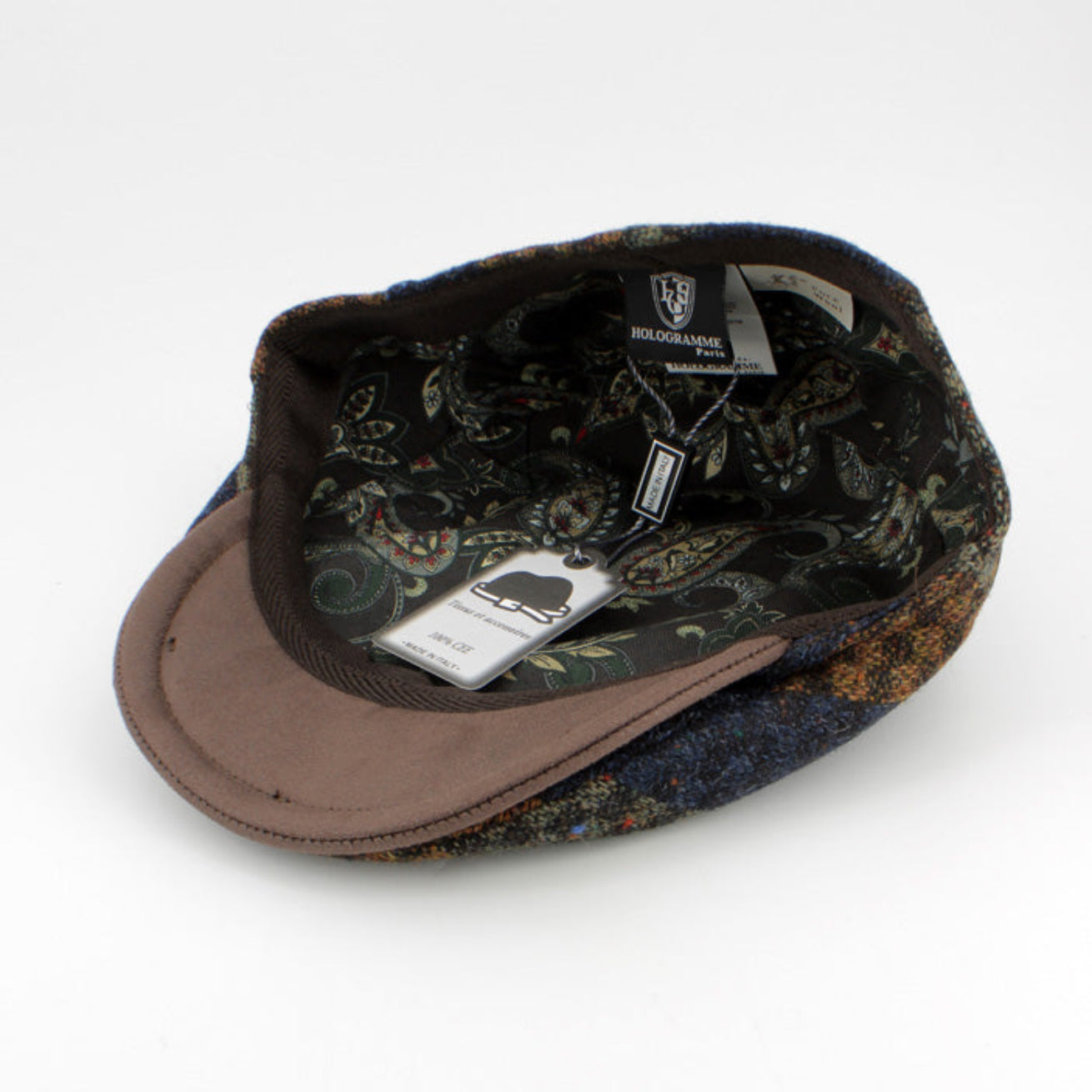 A vintage-style flat cap shown from an angled top-down view on a white background. The cap features a dark, ornate paisley patterned tweed exterior in black, blue, and brown tones, a solid brown fabric visor, and a dark patterned fabric lining with visible brand tags.