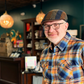 A smiling man wearing a brown and blue patterned flat cap, glasses, and a blue, orange, and yellow plaid button-down shirt, standing inside a shop with wooden shelves and hanging pendant lights in the background.