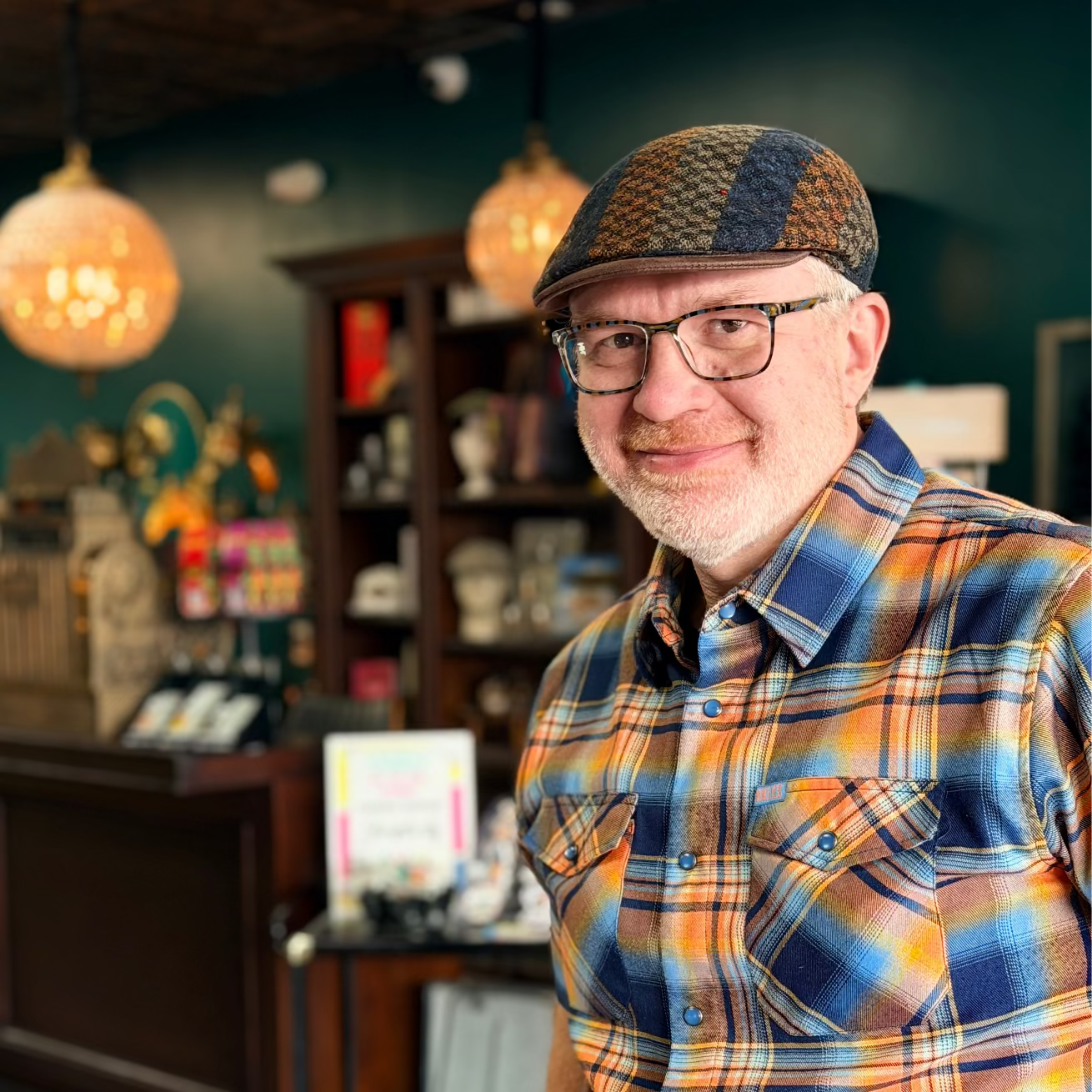 A smiling man wearing a brown and blue patterned flat cap, glasses, and a blue, orange, and yellow plaid button-down shirt, standing inside a shop with wooden shelves and hanging pendant lights in the background.