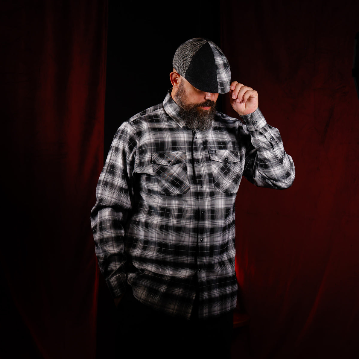 A man wearing a black and white plaid button-down shirt and a matching black and white plaid flat cap, posing against a dark red curtain backdrop.