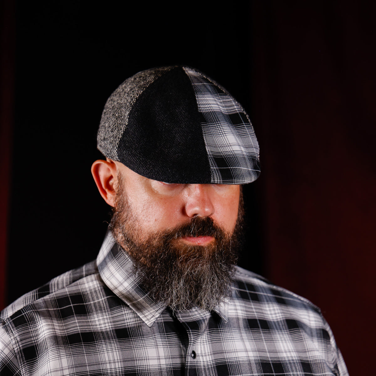 A man with a full beard wearing a black and gray plaid flat cap and a matching black and gray plaid button-down shirt, set against a dark background.