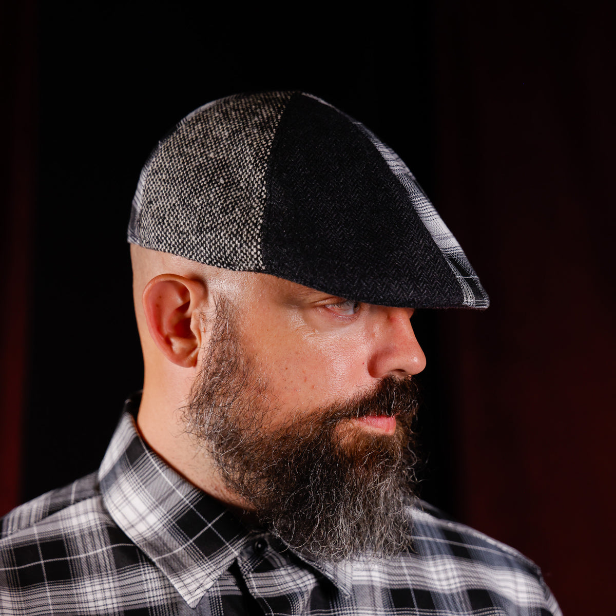 A man with a full beard wearing a black and gray herringbone flat cap and a black and white plaid shirt, shown in profile against a dark background.