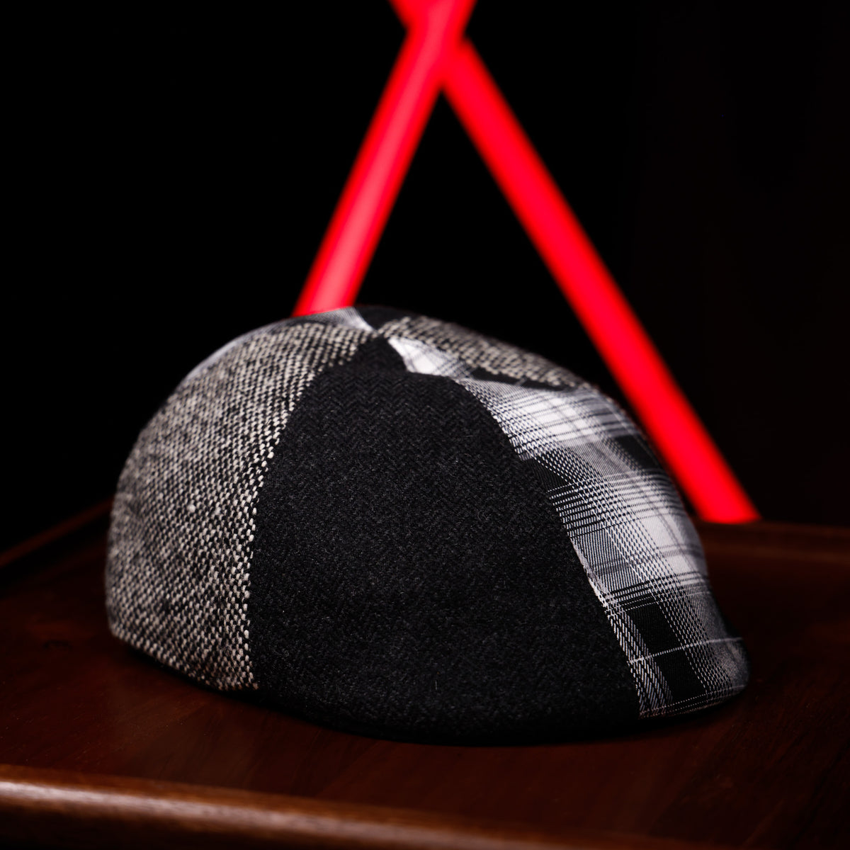 A black and gray plaid flat cap resting on a dark wooden surface with a black background and red geometric light beams behind it.