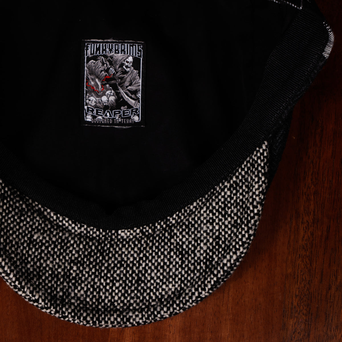 Close-up of a black trucker-style cap with a textured black-and-white brim, resting on a dark wooden surface. The inside of the crown features a black fabric lining with a rectangular embroidered patch showing a grim reaper and the text 'FUCK BEARZ' and 'REAPER OF THE DEAD'.