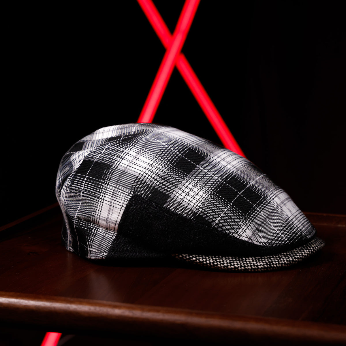 A black and white plaid flat cap with a black mesh brim, resting on a dark wooden surface with a red X-shaped light in the background.