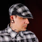 A man wearing a black, white, and gray plaid flat cap and a matching plaid button-down shirt, shown in profile against a dark studio background.