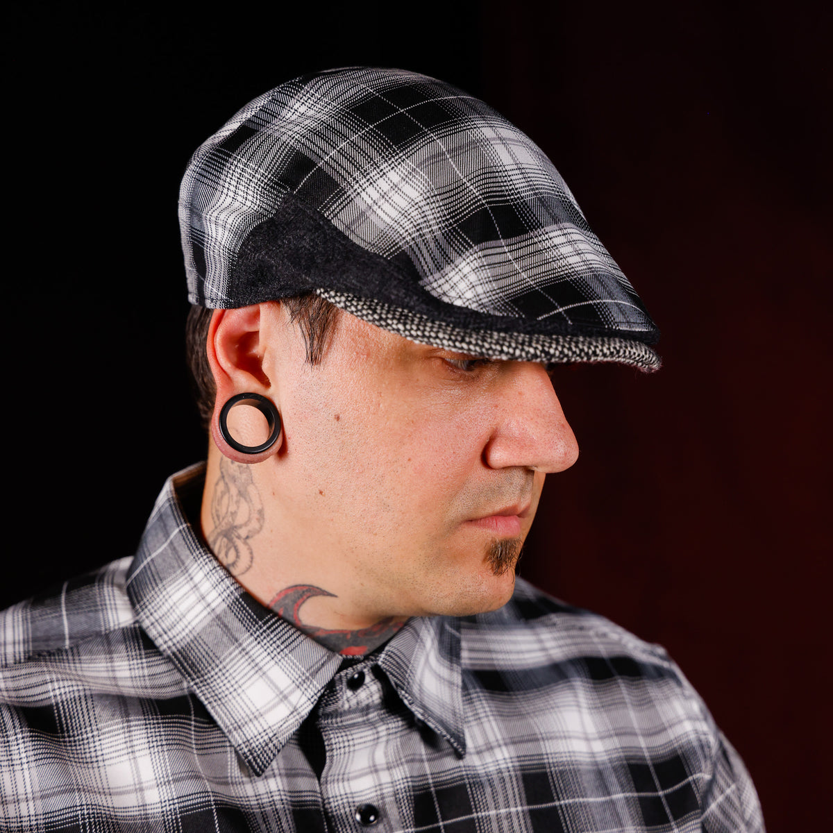 A man wearing a black, white, and gray plaid flat cap and a matching plaid button-down shirt, shown in profile against a dark studio background.