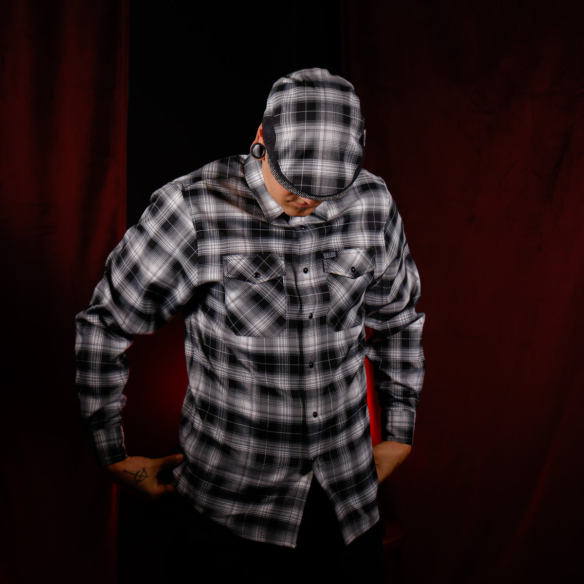 A man wearing a black and white plaid button-up shirt with matching plaid baseball cap, posed against a dark red curtain background.