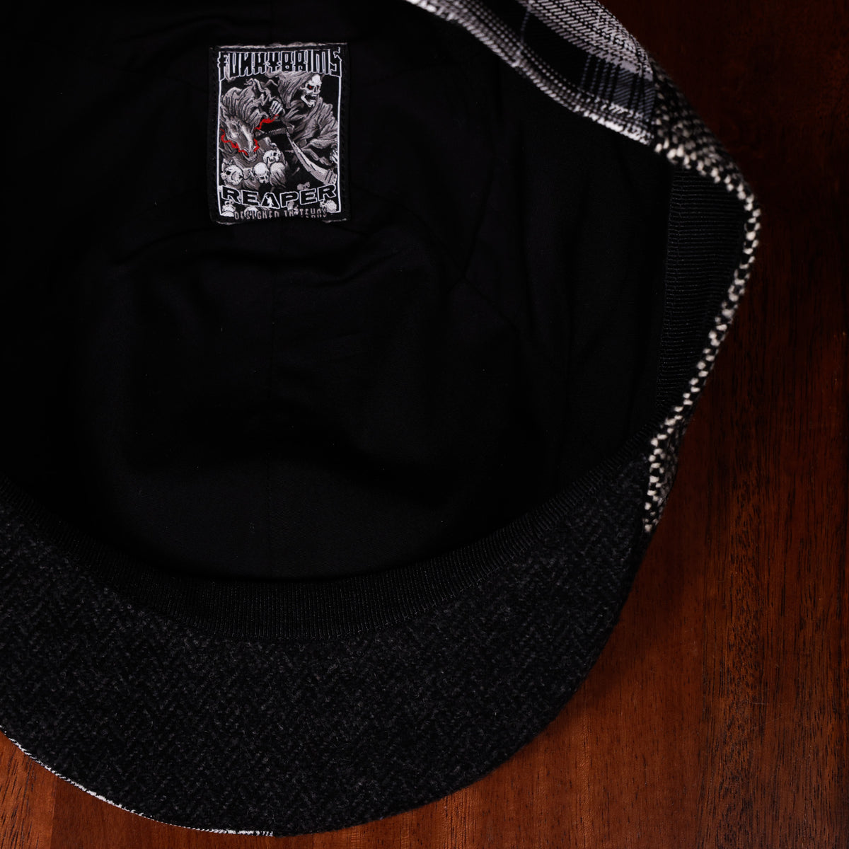 Close-up of the inside of a black cap resting on a dark wood surface, showing a printed graphic patch on the inner crown and a plaid-patterned lining along the edge.