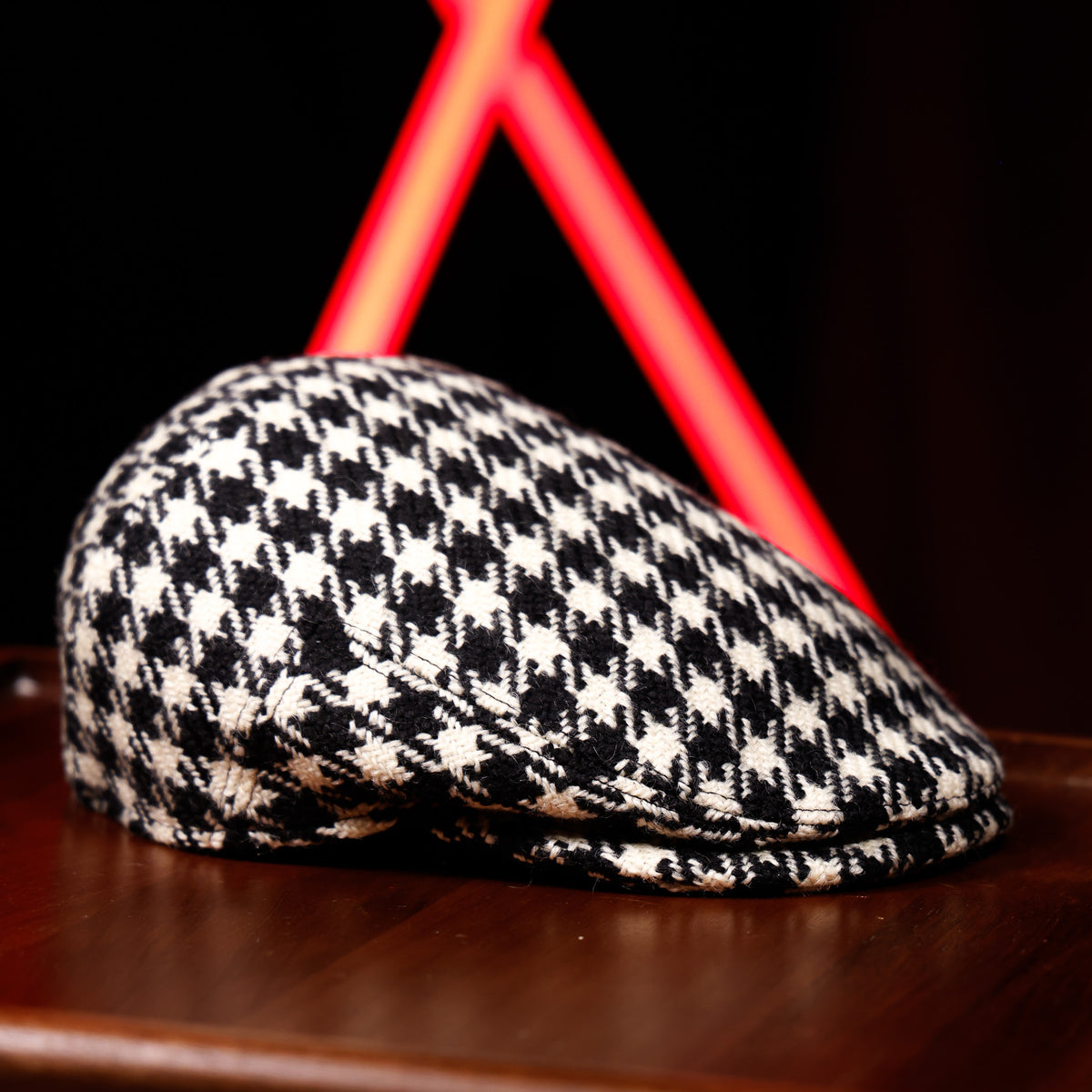 A black and white houndstooth patterned flat cap resting on a dark wooden surface, with a blurred red neon light in the background.
