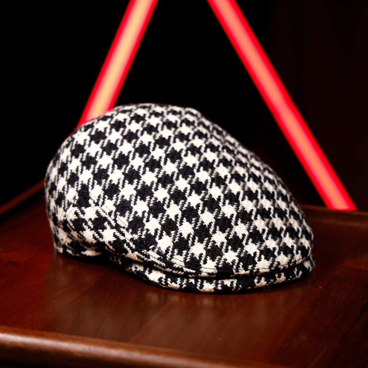 A black and white houndstooth patterned flat cap resting on a dark wooden surface, with a blurred red neon light in the background.