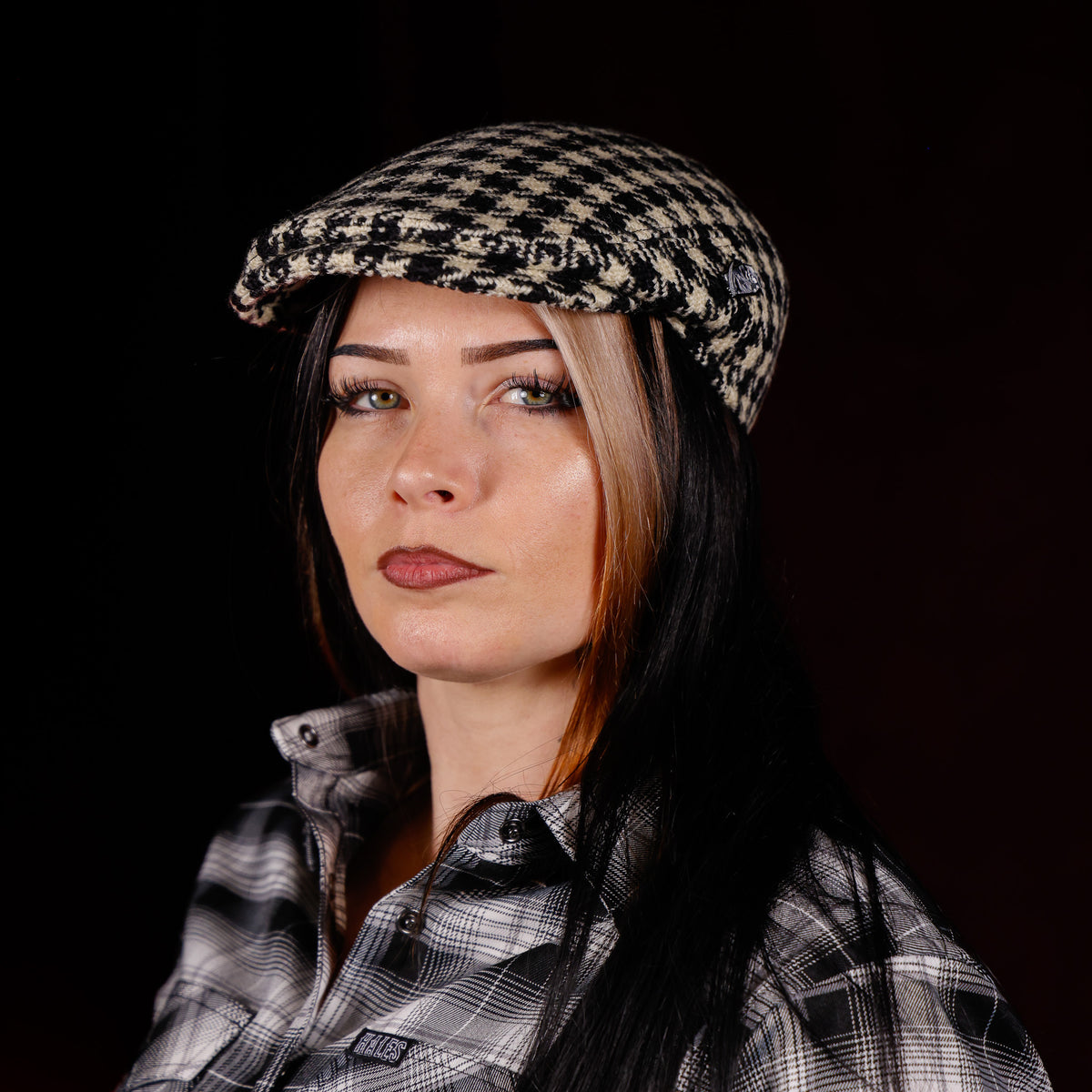 A woman with long dark hair and a blonde streak wearing a black and beige houndstooth flat cap and a matching plaid shirt, photographed against a solid black background.