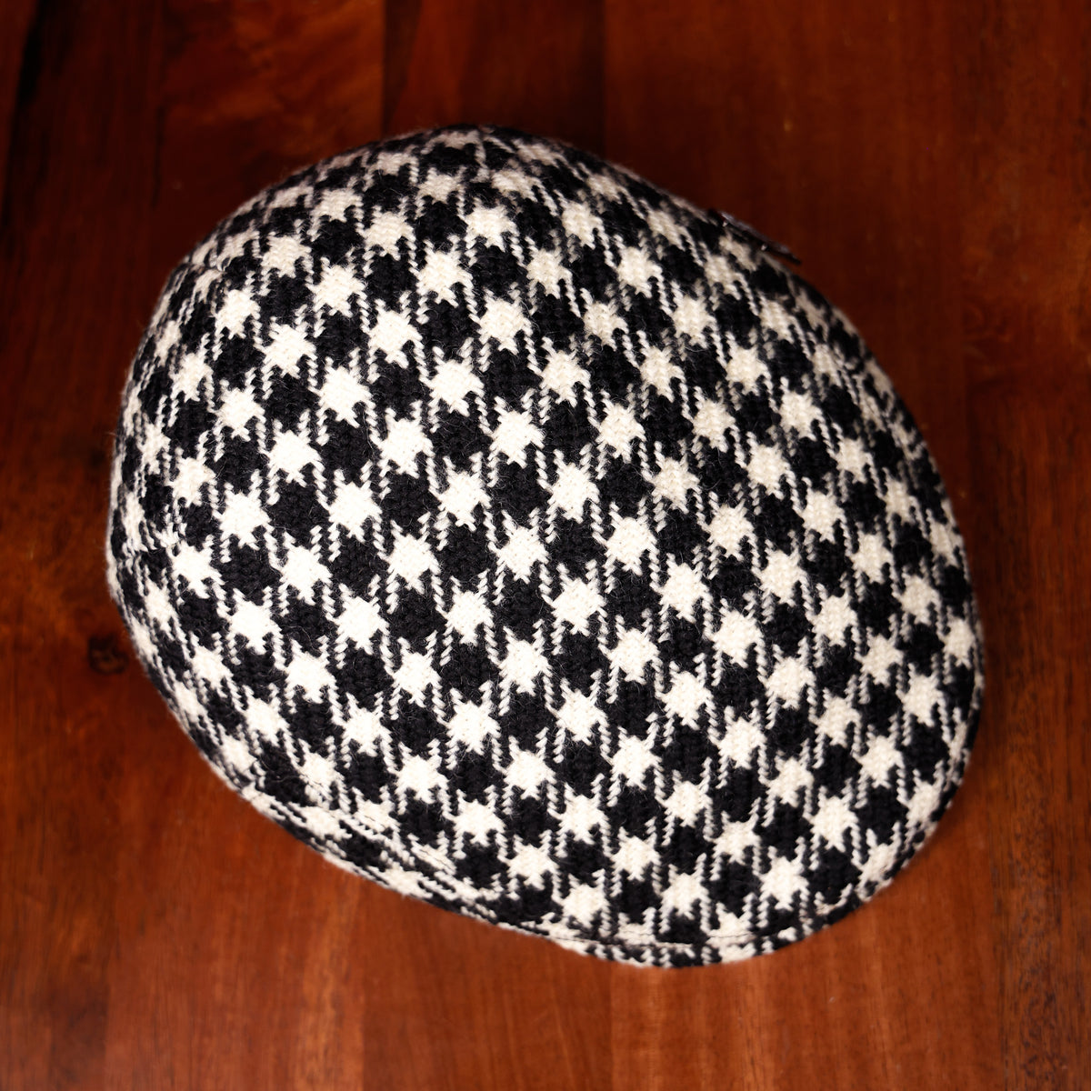 A top view of a black and white houndstooth patterned flat cap resting on a dark wooden surface.