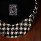 A close-up of a black and cream houndstooth patterned scally cap resting on a dark wood surface, showing the black fabric interior with a FunkyBrims 'Reaper' patch.