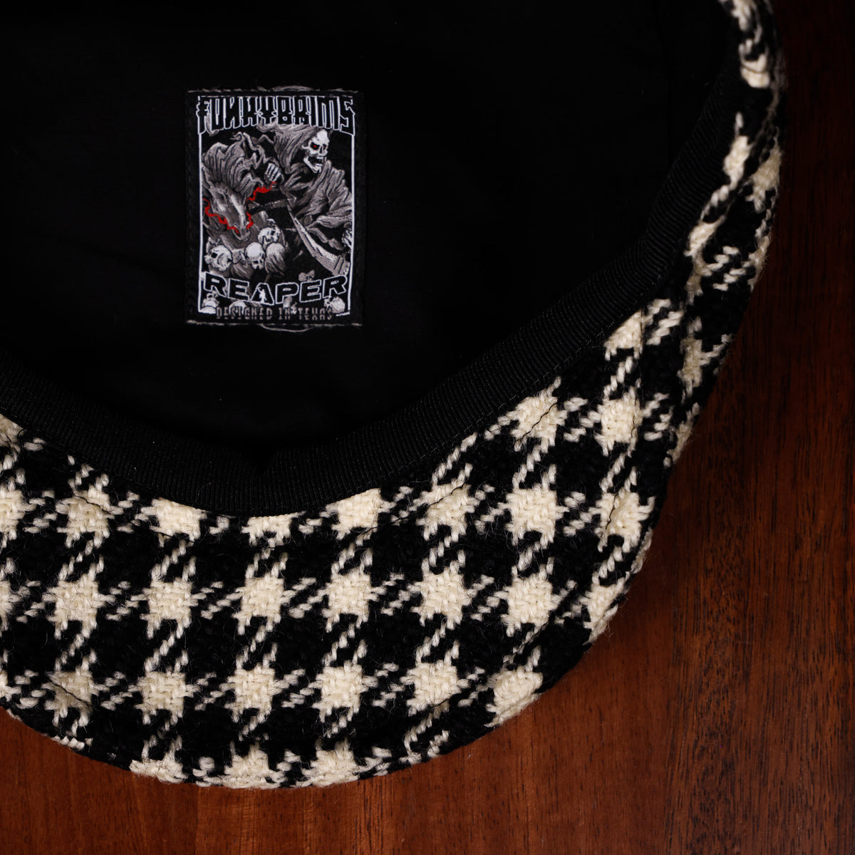 A close-up of a black and cream houndstooth patterned scally cap resting on a dark wood surface, showing the black fabric interior with a FunkyBrims 'Reaper' patch.