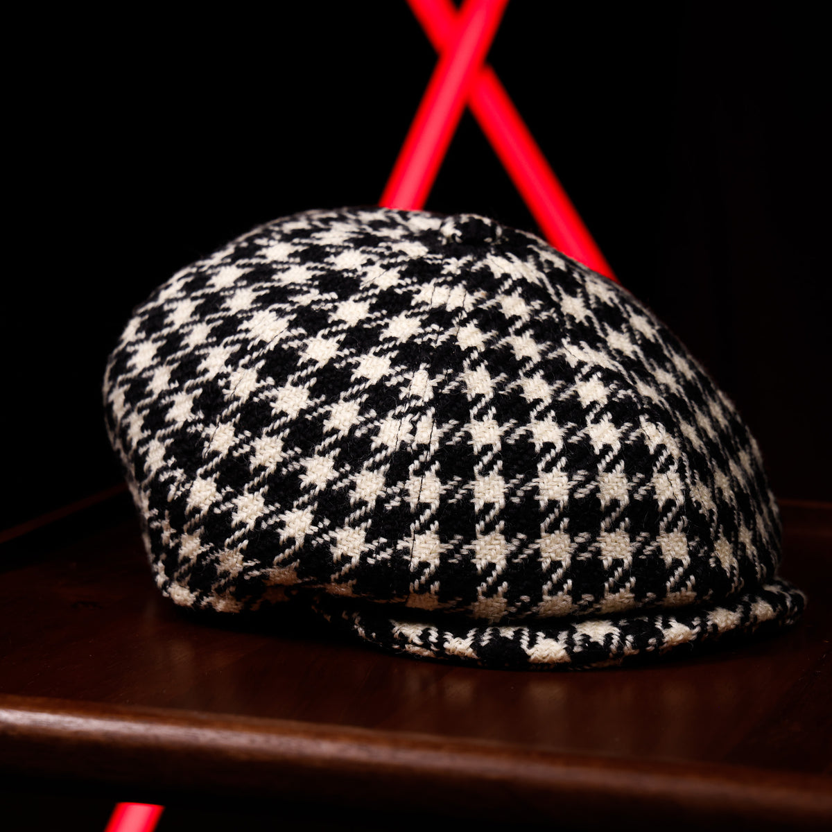 A black and white houndstooth flat cap resting on a dark wooden surface, with a blurred black background and bright red geometric light accents behind it.