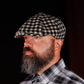 A man with a full beard wearing a black and beige houndstooth flat cap and a black and white plaid shirt, shown in profile against a dark background.