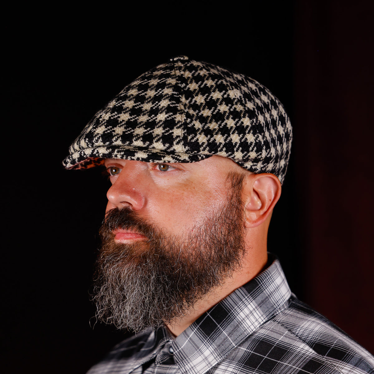 A man with a full beard wearing a black and beige houndstooth flat cap and a black and white plaid shirt, shown in profile against a dark background.