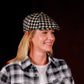 A woman wearing a black and beige houndstooth flat cap and a matching black and beige plaid button-up shirt, shown against a dark studio background.