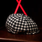 A black and off-white houndstooth flat cap resting on a dark wooden surface, with a blurred black background and bright red geometric lights behind it.