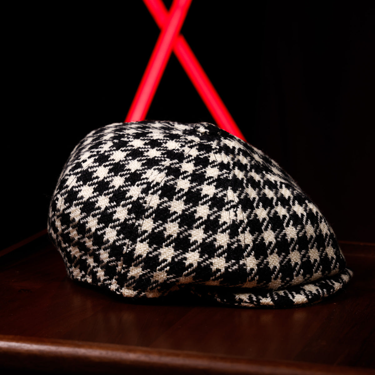 A black and off-white houndstooth flat cap resting on a dark wooden surface, with a blurred black background and bright red geometric lights behind it.