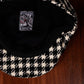 A close-up of a black and cream houndstooth patterned cap laid flat on a dark wood surface, showing the black interior and a sewn-in patch with the brand name FunkyBrims with Reaper.