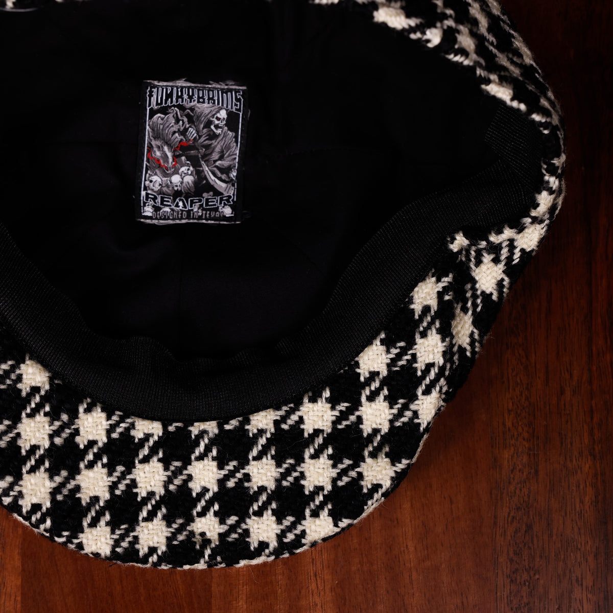 A close-up of a black and cream houndstooth patterned cap laid flat on a dark wood surface, showing the black interior and a sewn-in patch with the brand name FunkyBrims with Reaper.