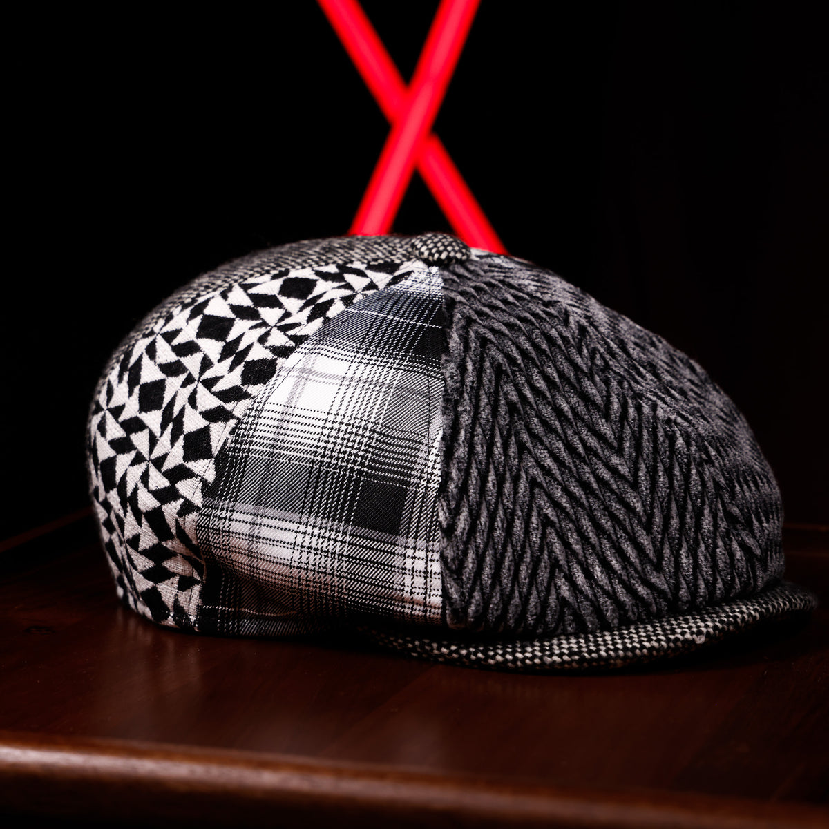 A structured flat-brim cap with three distinct patterned panels: a black and white geometric print, a black and white plaid, and a black and white textured herringbone weave. The cap is displayed on a dark wooden surface with a black background and a red neon light in the background.