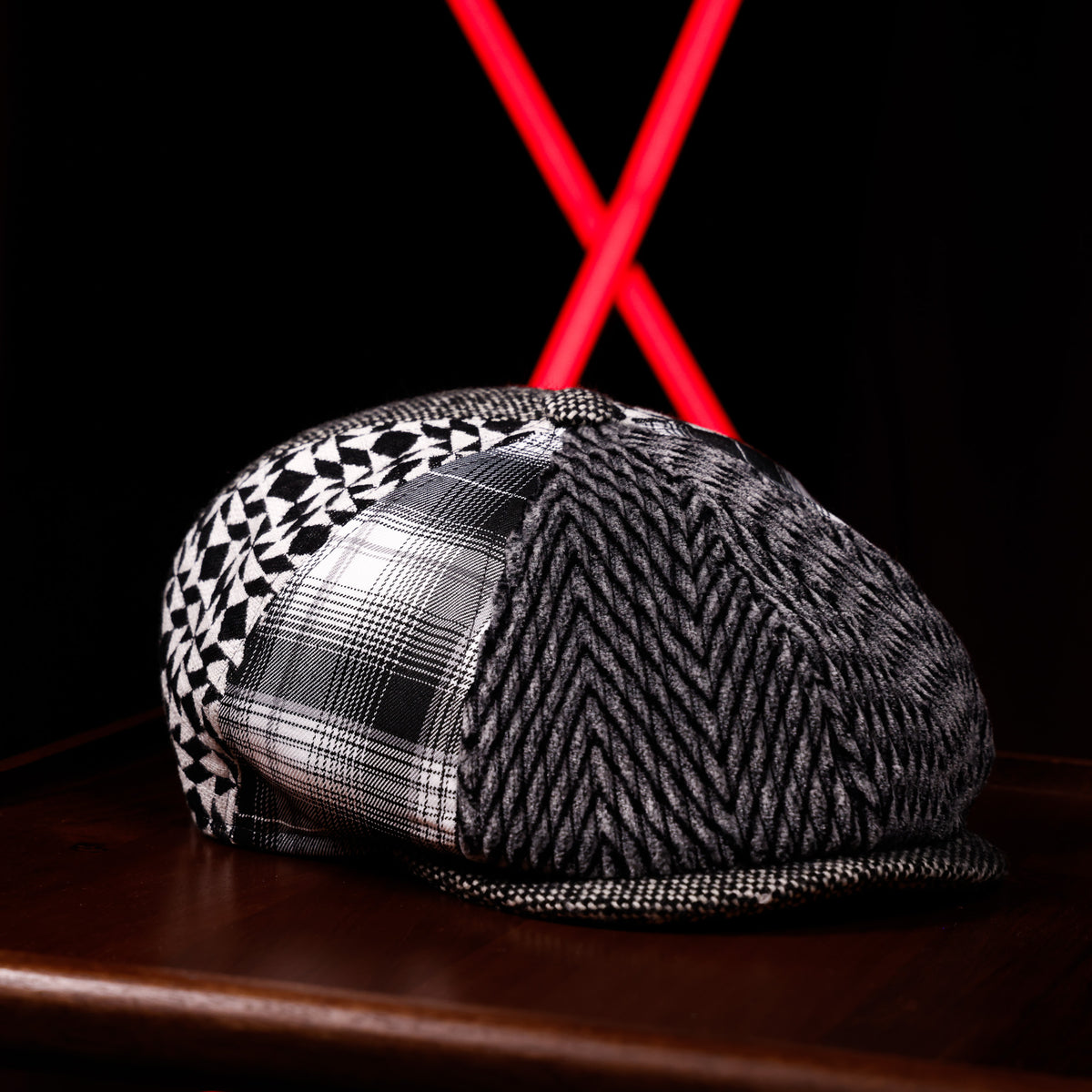 A structured flat-brim cap with a black and white herringbone pattern on the front, a black and white geometric pattern on the side, and a black and white plaid panel on the other side. The cap is resting on a dark wooden surface with a black background and a red X-shaped light in the distance.