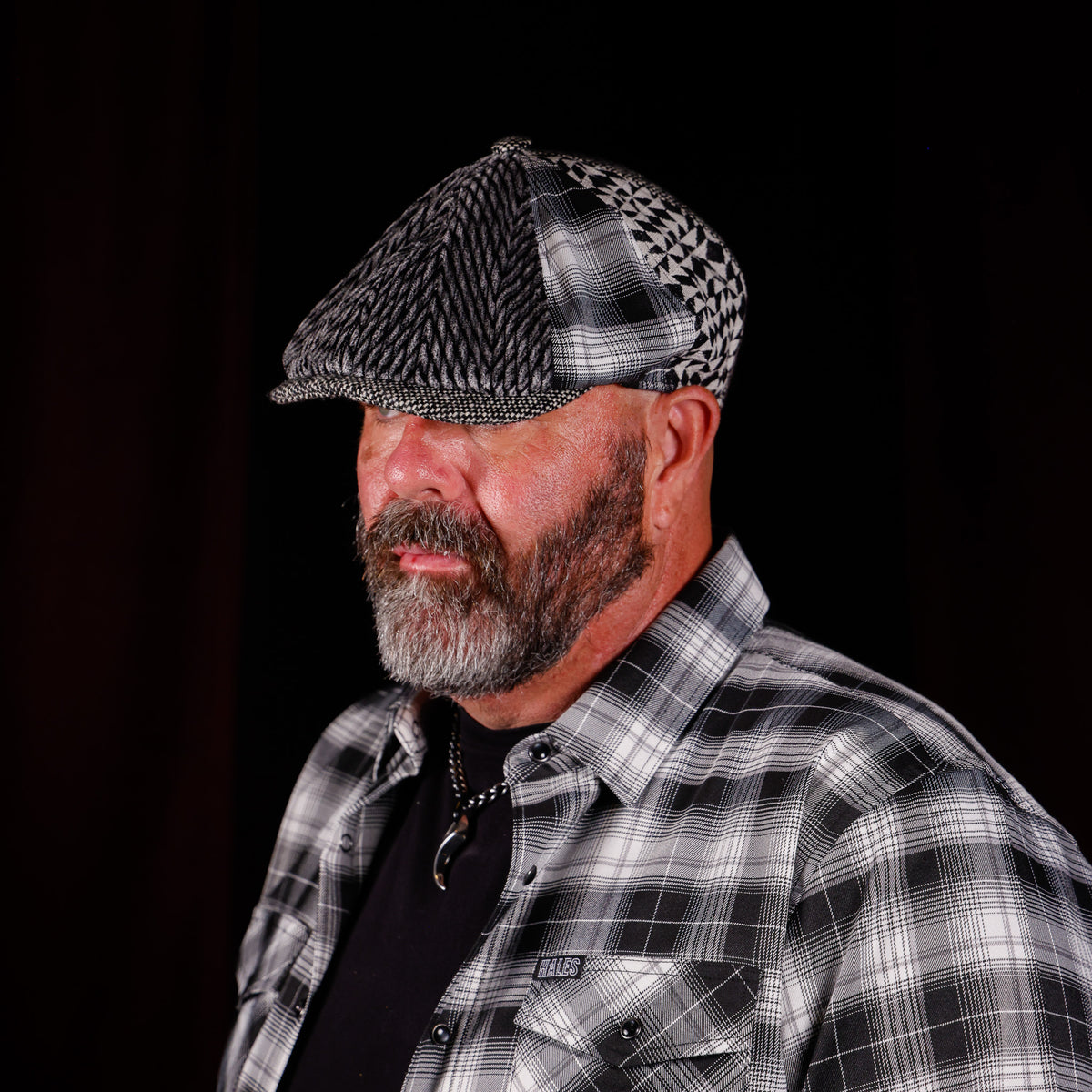 A man with a gray beard wearing a black and white patterned flat cap and a matching black and white plaid button-down shirt over a black t-shirt, photographed against a solid black background.