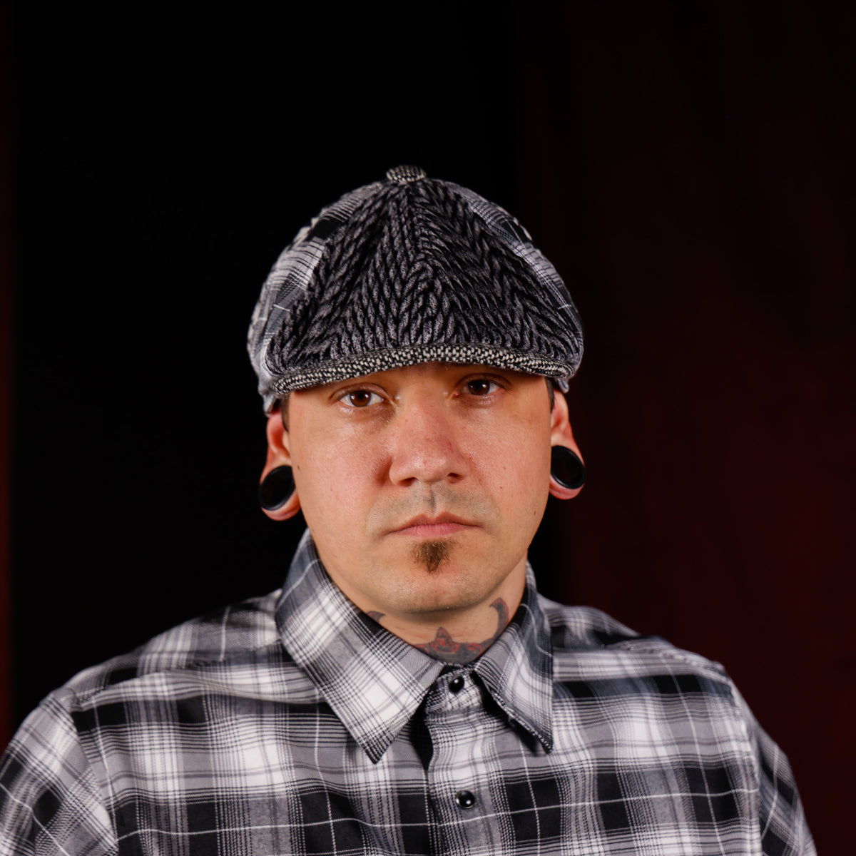 A man wearing a black and white plaid flat cap and a matching plaid button-down shirt, with large black ear cuffs, photographed against a dark studio background.