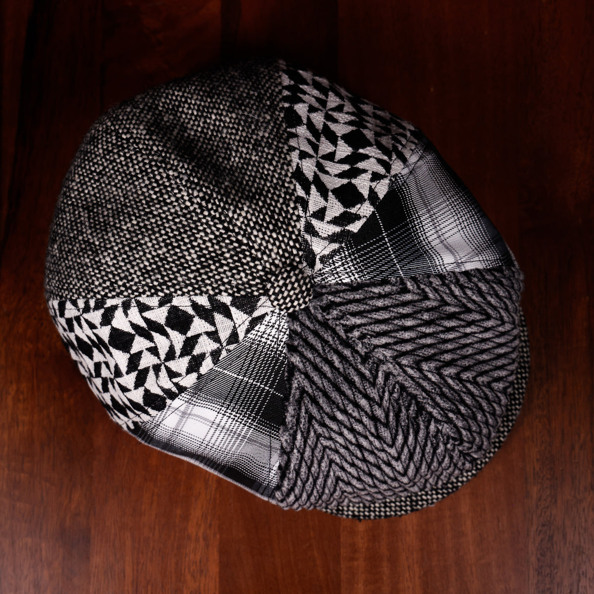 A patchwork flat cap made from multiple fabrics including black and white geometric patterns, plaid, and herringbone, photographed from a top-down angle on a dark wood background.