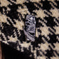 A coffin-shaped enamel pin featuring a black and white illustration of the Grim Reaper, resting on a black and cream houndstooth patterned fabric background.