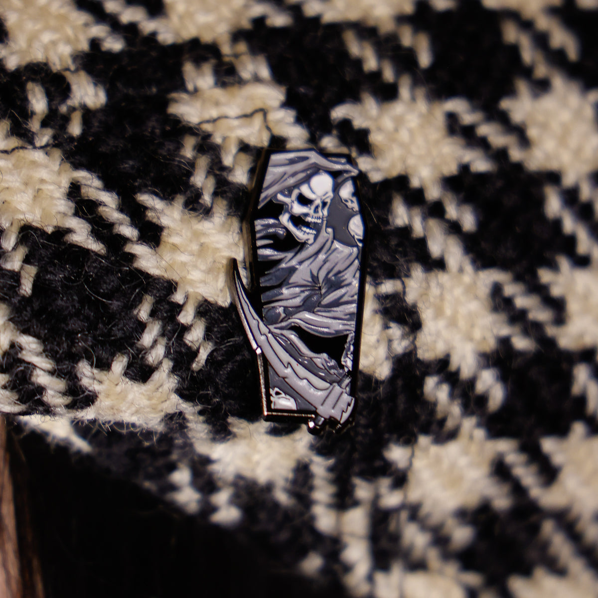 A coffin-shaped enamel pin featuring a black and white illustration of the Grim Reaper, resting on a black and cream houndstooth patterned fabric background.