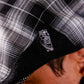 A close-up of a person wearing a black and white plaid cap, focusing on a small silver metal pin shaped like a tombstone with a skeleton design on the black brim.