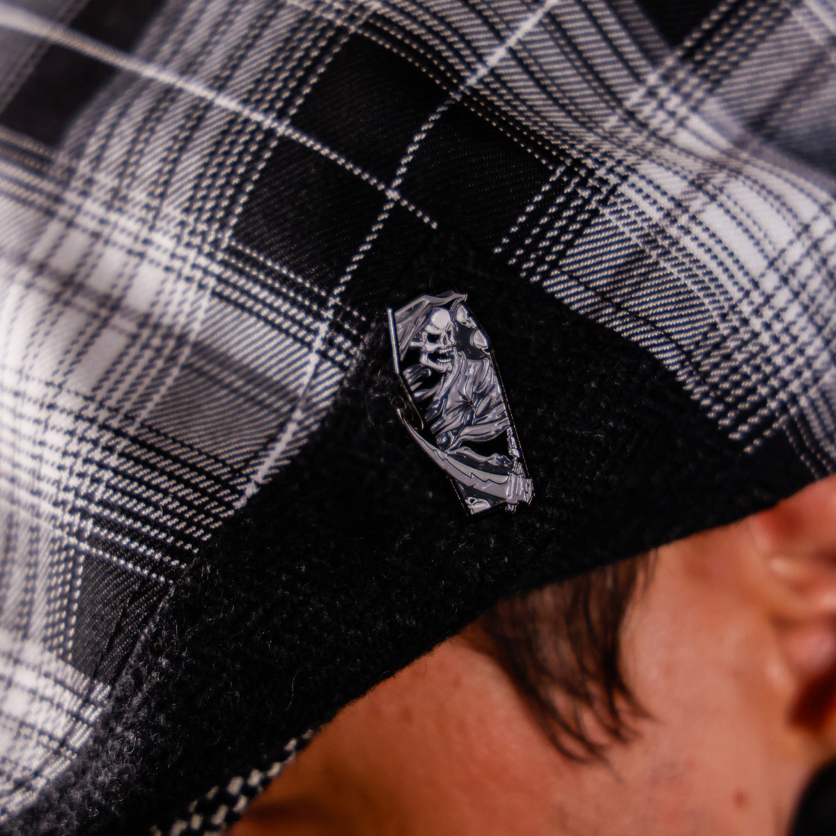 A close-up of a person wearing a black and white plaid cap, focusing on a small silver metal pin shaped like a tombstone with a skeleton design on the black brim.