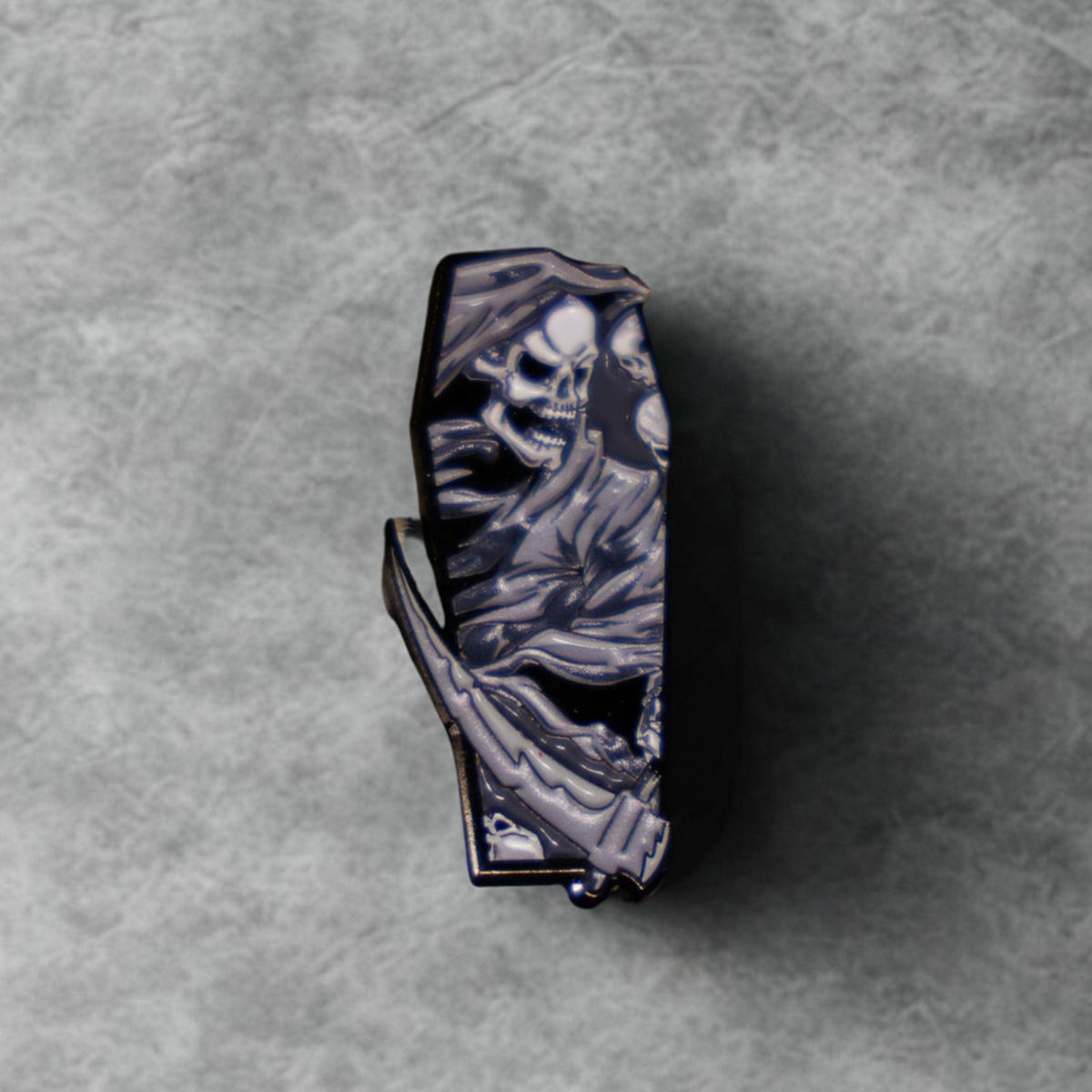 A coffin-shaped lighter with a detailed grim reaper design, featuring a metallic silver and black finish, set against a textured gray background.