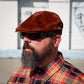 A man with a full beard wearing a brown suede flat cap, black sunglasses, and a colorful orange, yellow, and black plaid button-down shirt, photographed outdoors against a white wall with painted faces.