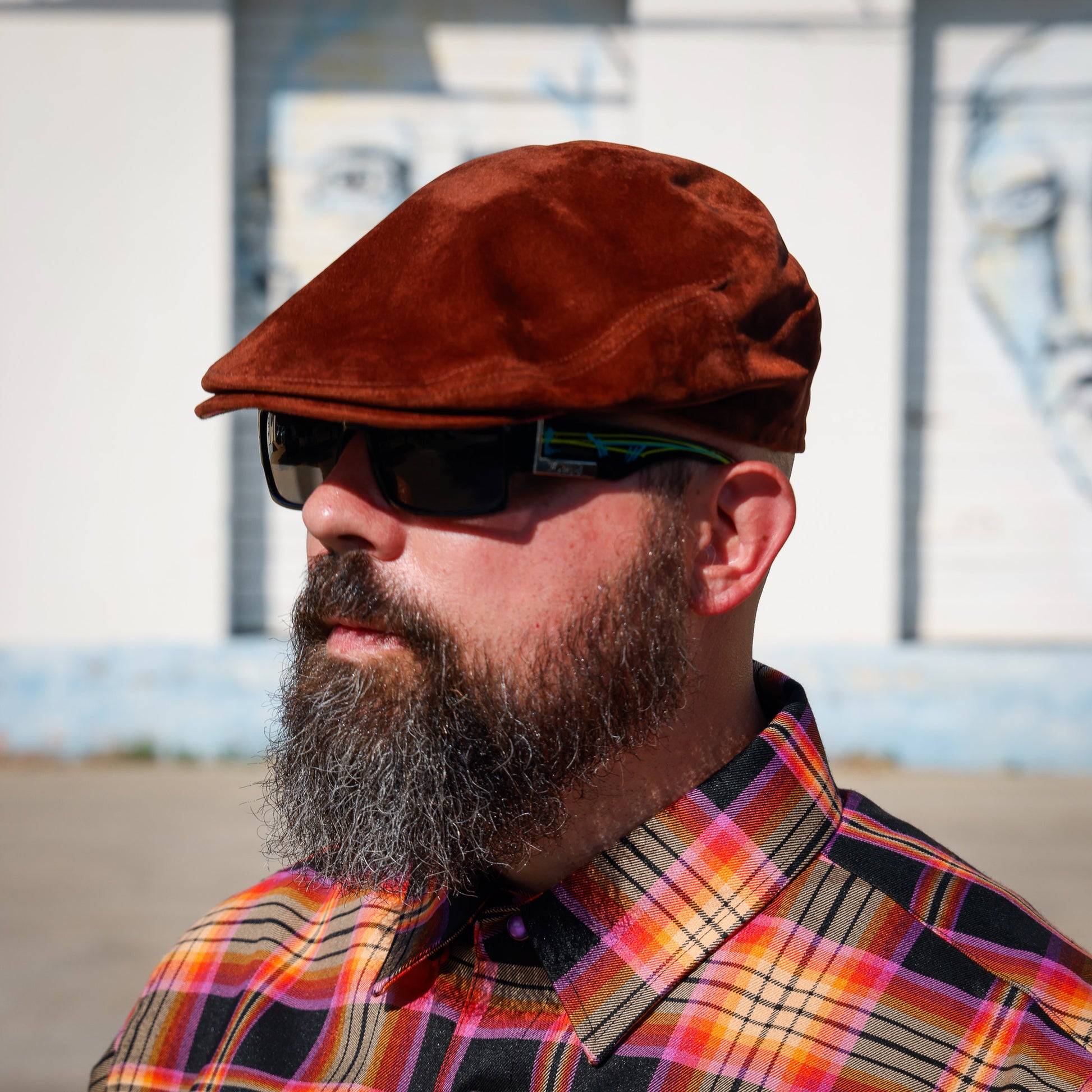 A man with a full beard wearing a brown suede flat cap, black sunglasses, and a colorful orange, yellow, and black plaid button-down shirt, photographed outdoors against a white wall with painted faces.
