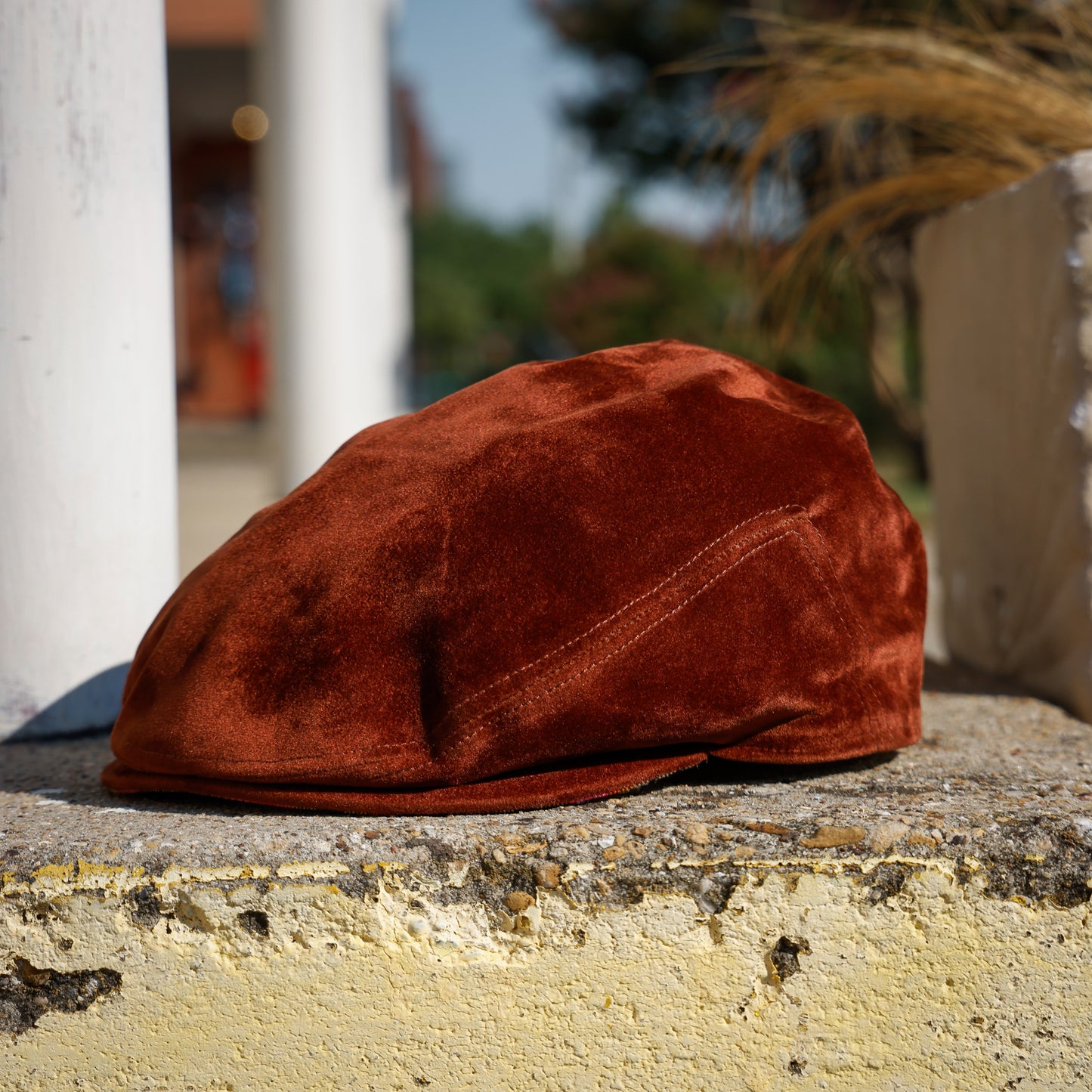 A rust-brown velvet flat cap resting on a weathered stone ledge outdoors, with blurred columns and greenery in the background.