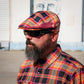 A man with a full beard and sunglasses wearing a colorful plaid flat cap and matching plaid button-down shirt, photographed outdoors against a light-colored building wall.