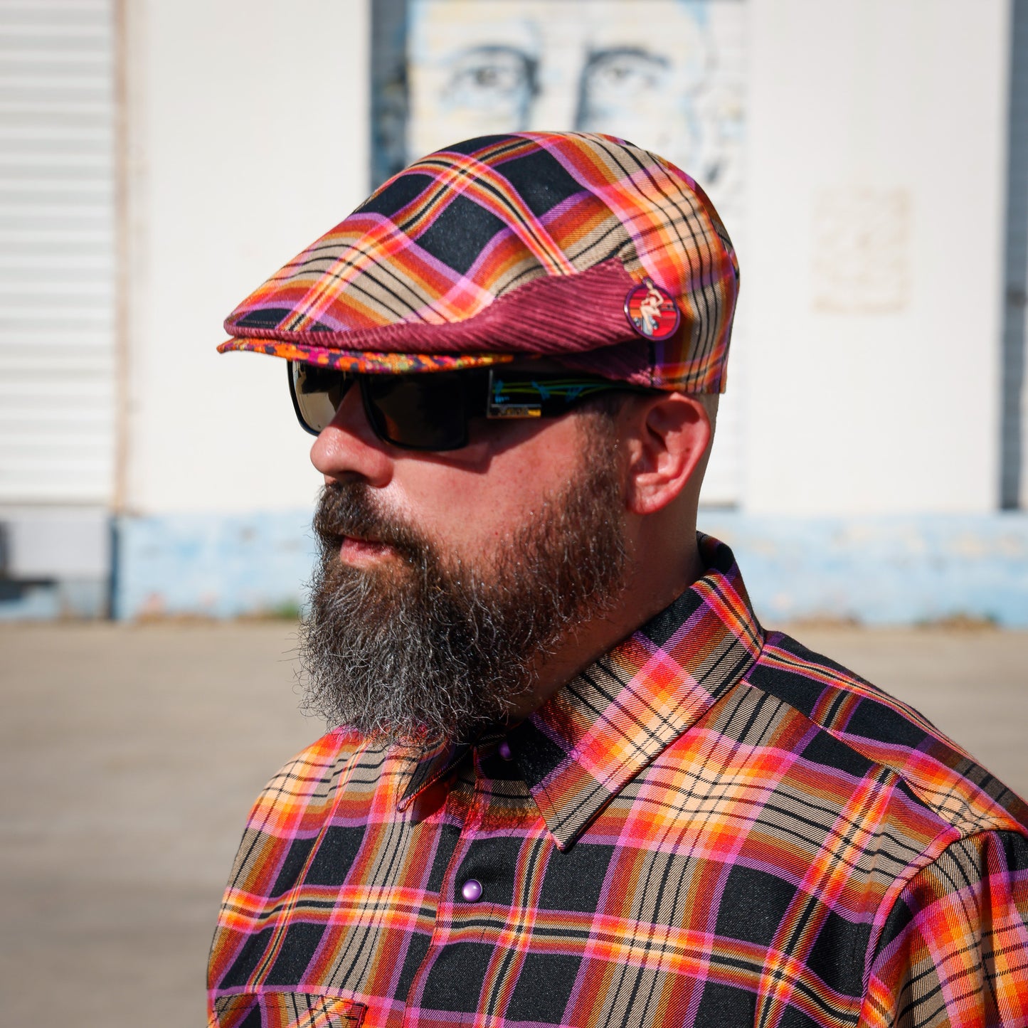 A man with a full beard and sunglasses wearing a colorful plaid flat cap and matching plaid button-down shirt, photographed outdoors against a light-colored building wall.