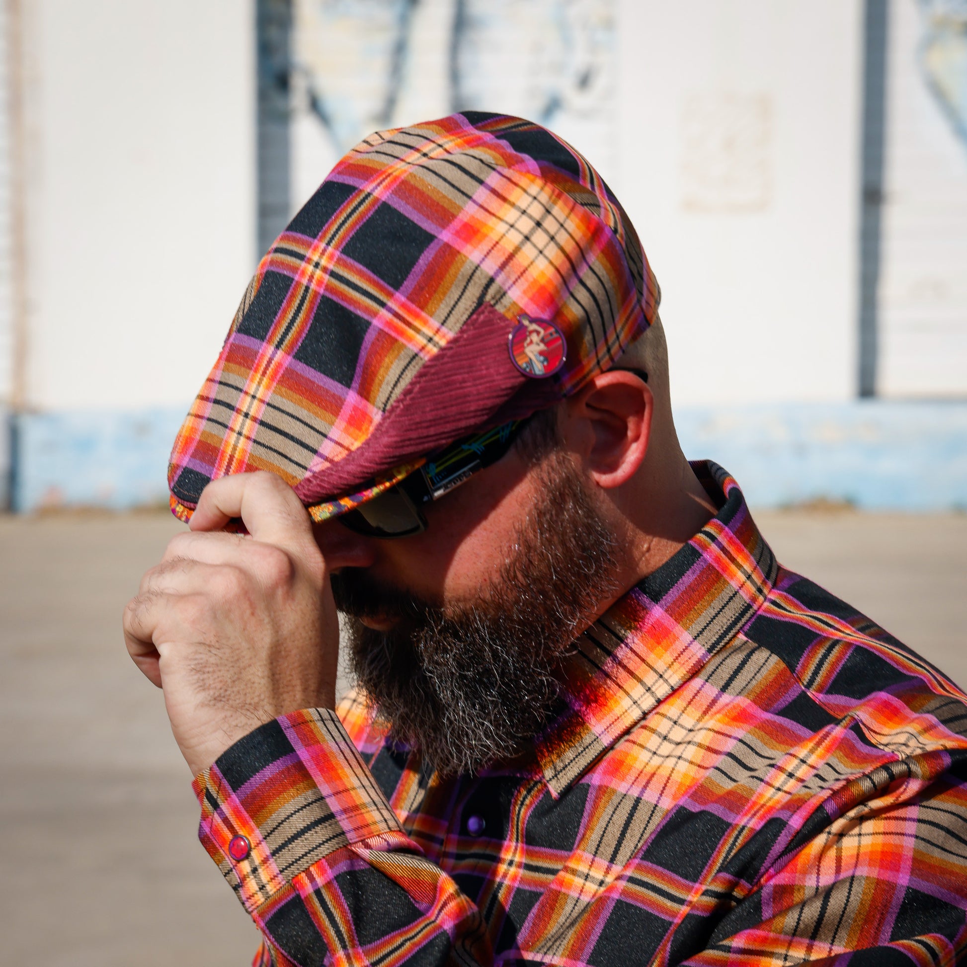 A man wearing a colorful plaid flat cap and matching plaid button-down shirt, holding his cap in front of his face. The plaid pattern features orange, yellow, pink, and black stripes.