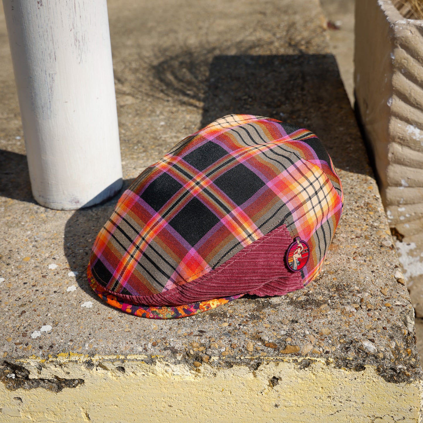 A plaid flat cap with orange, pink, black, and beige stripes, featuring a burgundy corduroy visor and a small embroidered logo patch, resting on a concrete ledge outdoors.