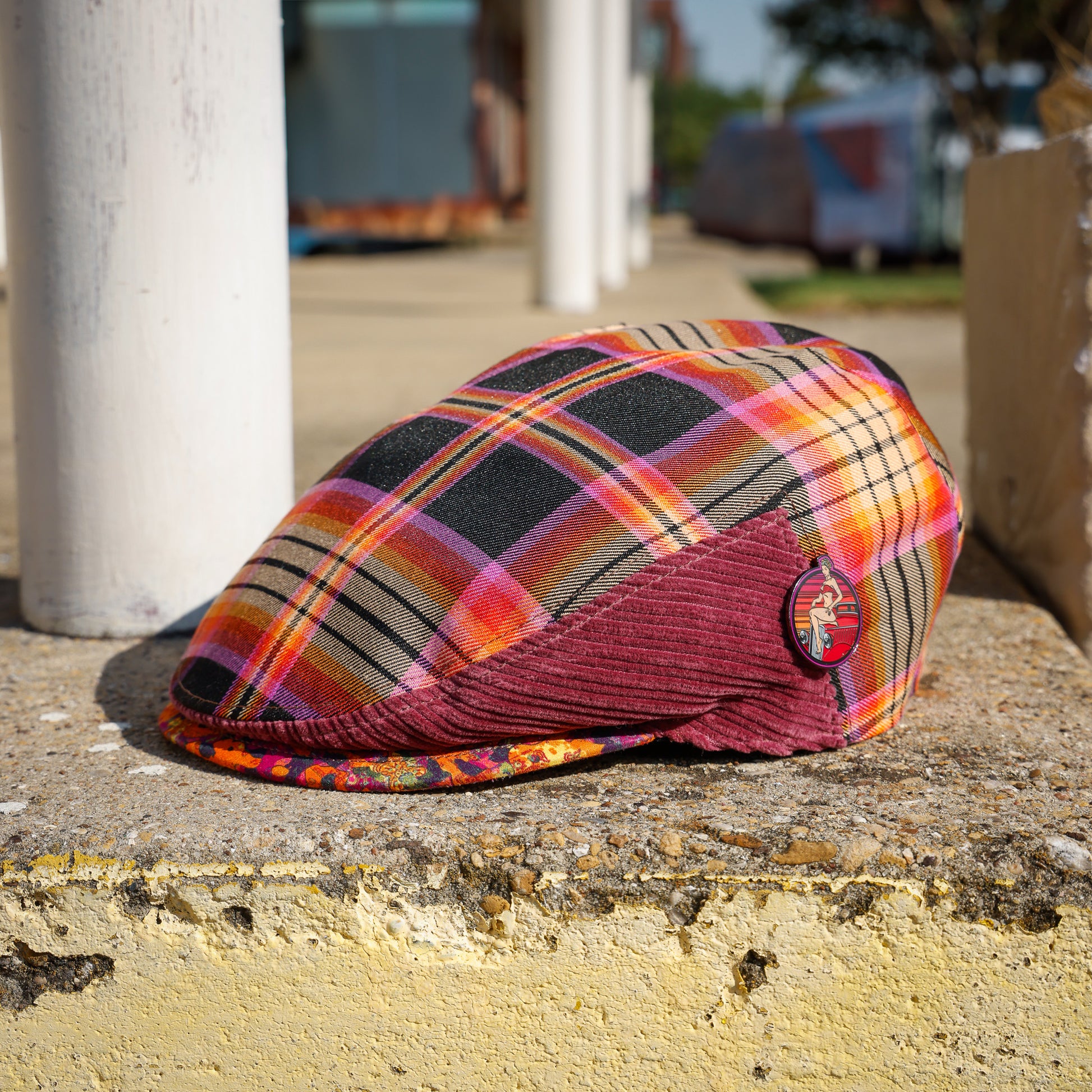 A plaid flat cap with a burgundy corduroy visor sits on a weathered concrete ledge outdoors. The cap features a multicolor plaid pattern with black, orange, pink, and yellow tones, and a small circular badge is attached to the side.