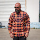 A man wearing a long-sleeve button-down plaid shirt in red, orange, yellow, and black, standing outdoors in front of a white metal garage door.