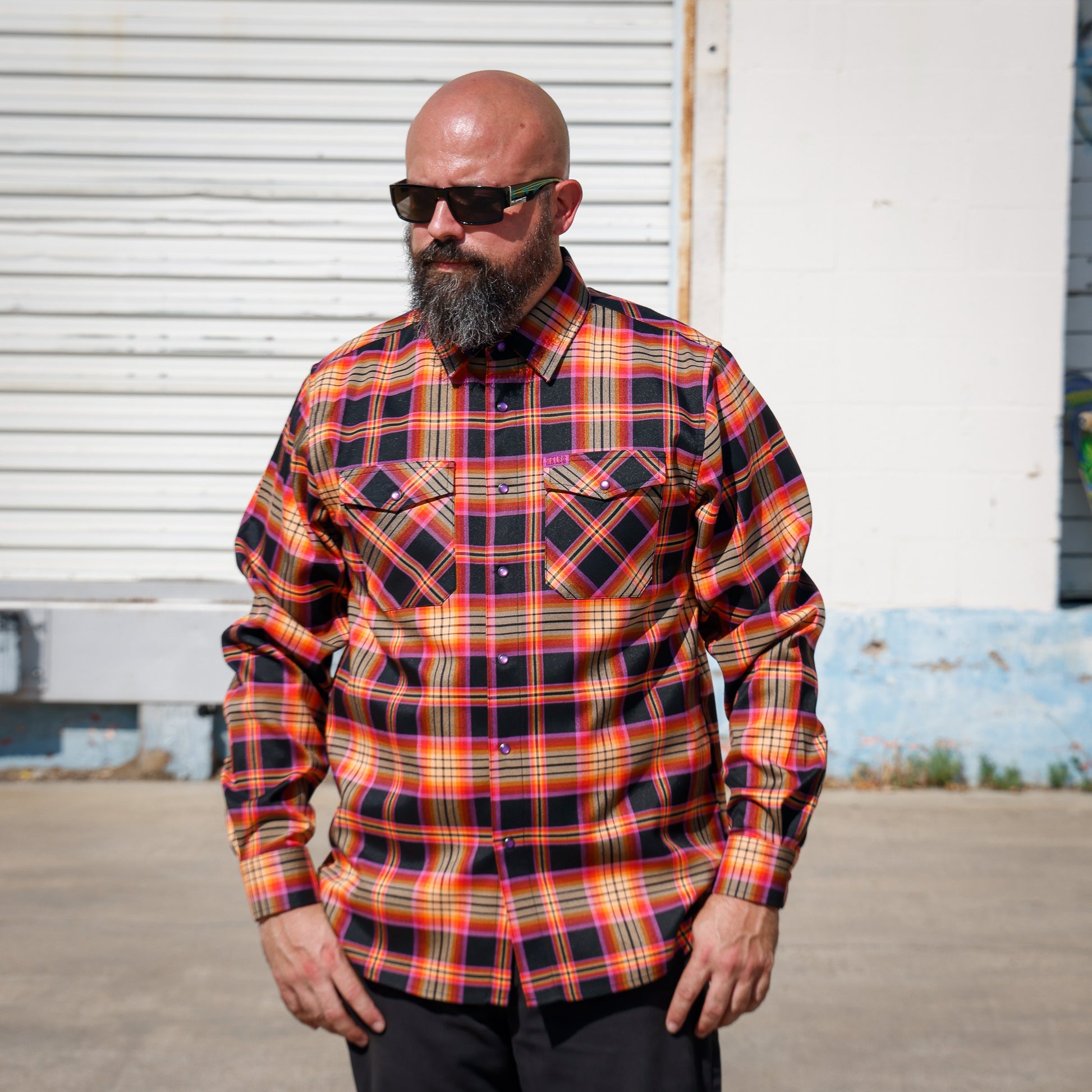 A man wearing a long-sleeve button-down plaid shirt in red, orange, yellow, and black, standing outdoors in front of a white metal garage door.