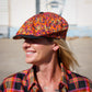 A woman wearing a vibrant orange and purple patterned flat cap and a matching orange and purple plaid shirt, photographed outdoors in bright sunlight.